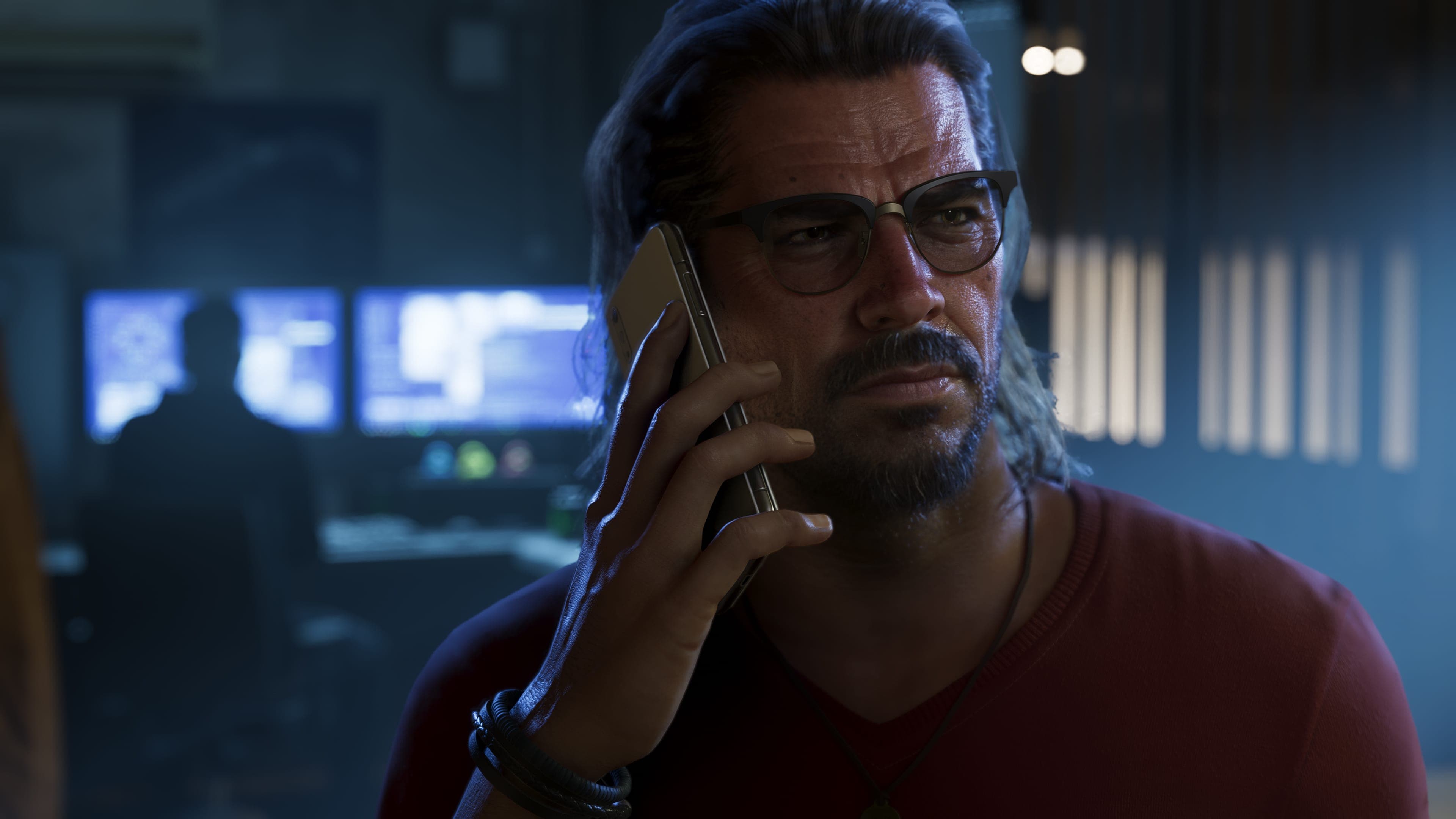 Official Rockstar Games render of Raul Bautista — a seasoned bank robber, depicted on a phone call in a dim interior, wearing glasses and a red sweater. Computer monitors with surveillance feeds are visible in the background.